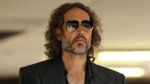 A headshot of Russell Brand who wears a suit and sunglasses.