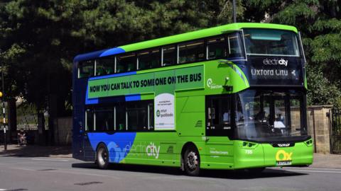 A green electric Oxford City bus on a street. The words 'Now you can talk to someone on the bus' are printed on the side of the vehicle. It is a sunny day.