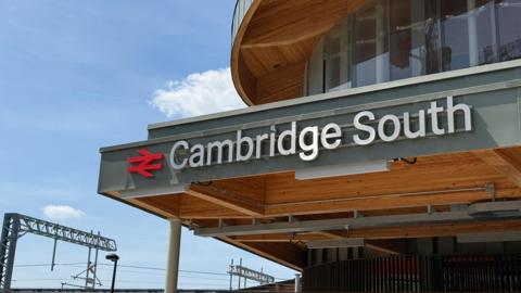 A train station's entrance has a sign above it that says Cambridge South. There are overhead cables on the left where a train line runs. 