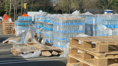 Stacks of bottled water set up in a car park, next to empty pallets.