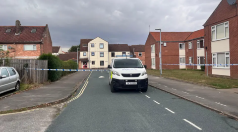 A road with a police van parked facing towards the camera. A blue and white police cordon is closing off the area. Houses are surrounding the road.