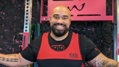 Ram Patten. He is bald and has black facial hair. He is wearing black and red gym clothing and is sat by some gym equipment. He has his arms out, showing his tattoos. Ram is looking directly at the camera and smiling 