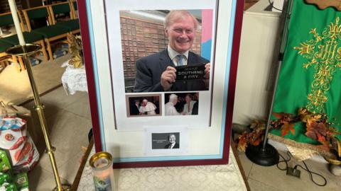 A frame holds images of the late MP. One shows him holding a flyer saying make Southend a city. the two others are of him meeting the Pope.