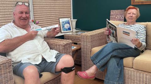 Peter and Pat sit on adjacent wicker chairs with cushions on them. He holds up his silver desk ID, which has his name on and the Barclaycard logo. Pat holds open a Barclays magazine with the car on the back. She has fair hair with an orange streak at the front.