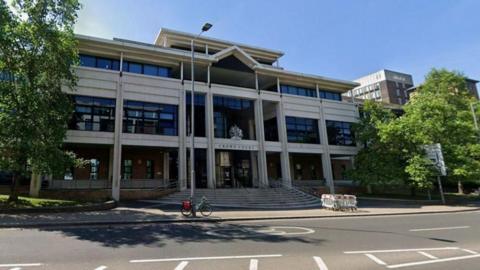 Kingston Crown Court - a grey, square building with trees on either side. There is a lamp post and a bike outside.