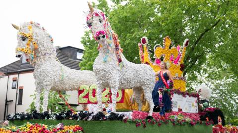 A float at the Spalding Flower Parade 2023 featuring two white unicorns with floral head accessories and a big golden crown and a woman with blonde hair and a red sash