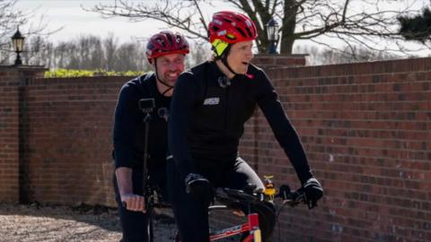 Greg James at the front of a tandem bike with Prince William sitting behind him. They both smile and wear black long-sleeve tops and red helmets.