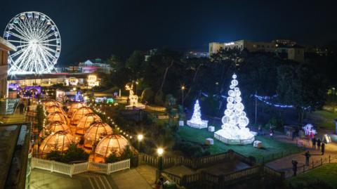 Two christmas trees made of white baubles are liton the grass on the right of the scene. On the left in the foreground are two rows of plastic see-through igloos for dinners and in the background is a large ferris wheel.