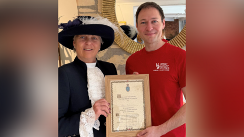 Adam is on the right wearing a red t-shirt with the Stand Against Violence logo and the female High Sheriff is on the left wearing her uniform of a dark blue jacket and white blouse with lacy collar and cuffs, and a dark blue-brimmed hat with a white and a blue feather