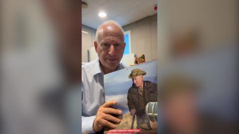 Steve Rosenberg holds a calendar featuring an image of Vladimir Putin wearing a green hat and brown jacket