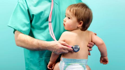 A nurse in green scrubs, holding a pink Stethoscope against the back of a young, brown haired toddler. The child is topless, wearing a nappy, facing the doctor and turning its head around 