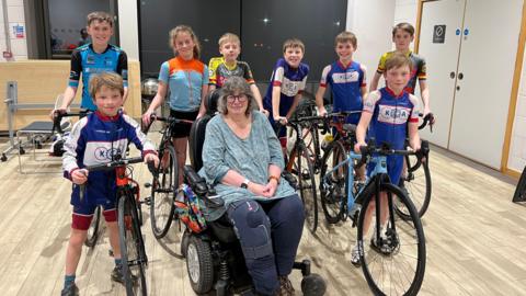 Six children in a assorted sports lycra stand holding bikes, in front of them are two boys also in cycling lycra holding bikes either side of a lady with round glasses and grey hair. She is in a wheelchair and is wearing a teal top and navy trousers, her right knee has a knee brace on it. 