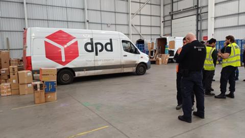 Men in high visibility jackets stood in a warehouse talking with a dpd van and parcels in the background.