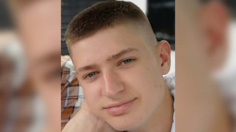 Alex Lewis is smiling while looking at the camera and resting his face on his right hand. He has blue eyes and short brown hair. 