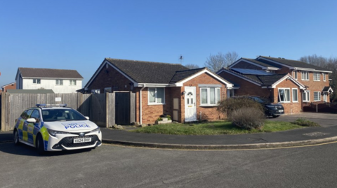 A police car parked outside a bungalow, with other houses alongside