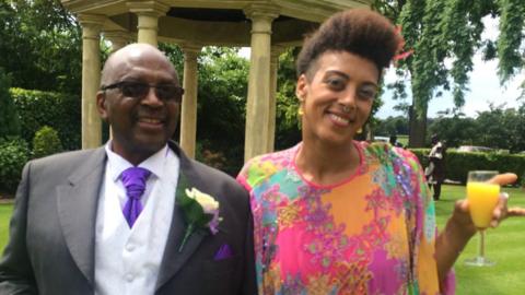 A father and daughter stand together for a photograph at a wedding. The father wears a grey formal suit with purple tie and rose boutonniere. He has a bald head and sunglasses and is smiling. The daughter wears a colourful dress, has a nose ring and is smiling. She has afro hair in a quiff style. They stand in front of a stone monument. 