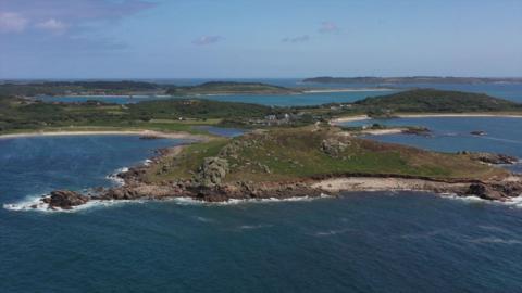 The island of Bryher is shown with rocky hills, sandy beaches and bodies of water beneath a blue sky scattered with clouds. 