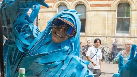 A man is dressed in an elaborate costume made of long strips of blue fabric ans is wearing large goggles. He is waving through a window at someone sitting inside. There are people walking on the street behind him