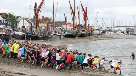 A group of about 100 people run down the mud into the River Chelmer estuary. Boats on the mudflats and buildings are in the background. 