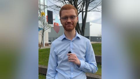 Scottish greens co-leader Ross Greer smiles at the camera as he stands in front of grass, a tree and a building. He has ginger hair, a ginger beard and is wearing glasses and a blue shirt.