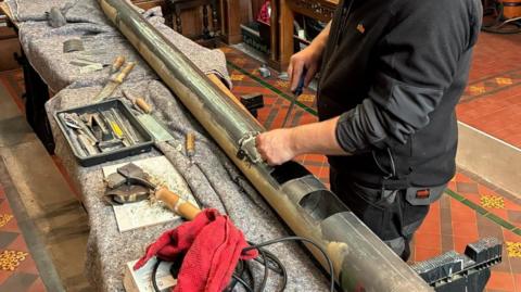 A large metal pipe, several metres long with open sections cut out, is lying on a table while someone works on it with a tool. The person's head is cropped out. Various other wooden handled tools are on the table.