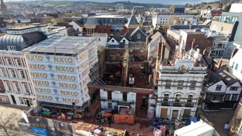 Aerial view of the Royal Clarence redevelopment site in Exeter, showing the fire-damaged former hotel under construction, with scaffolding, machinery and building materials visible.