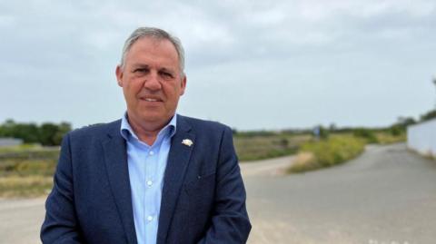 A man dressed in a dark blue blazer with a light blue shirt underneath. He has grey hair and a small broach on his blazer which is silver. He is stood in front of sand dunes and a road with grassy patches on the dunes.