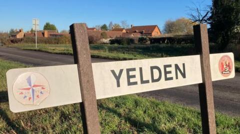 A village sign, saying Yelden, on capital letters, with two emblems either side of the sign. A village can be seen in the distance and grass below the sign.