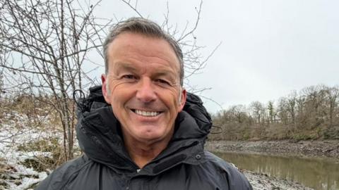 Gary Bankhead standing by the river. He has short, greying hair and is smiling at the camera. he is wearing a thick coat. The River Wear curves to the left behind him. Snow can be seen on the grass to the left.