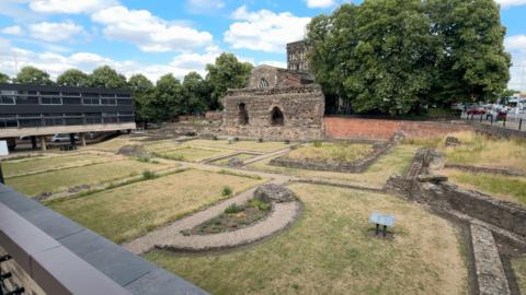 A view of a Roman site with grass in between stone passageways and an ancient wall at the back.