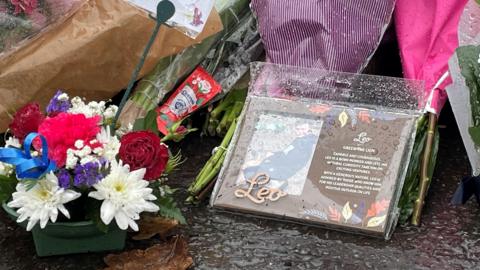 A close-up photograph of floral tributes. Among the flowers is a photo of the boy with the word 'Leo'  attached to it and what appears to be a poem.