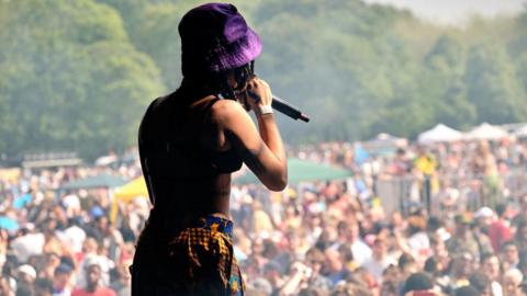 A female singer on stage wearing a purple hat, cropped black top and patterned yellow and blue trousers. She is singing into a microphone towards a large crowd.