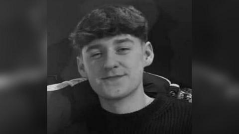 A young man in his late teens wearing a black jumper. He had dark hair and an arm is around his shoulder. The picture is monochrome.