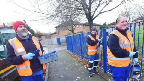 Three caretakers - two men and a woman - are painting a park fence blue. They are wearing blue work trousers and fleeces with high visibility orange vests on top as they hold cans of paint and brushes. 