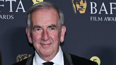 Robert Harris, wearing a black suit standing in front of an advertising banner for the Bafta Film Awards on Sunday