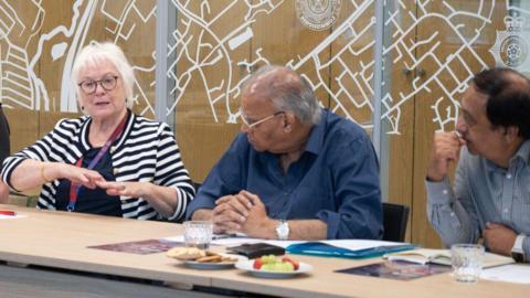 A woman in a striped top speaking to two men all sat behind a table. On the table are papers, fruit and biscuits and glasses of water.