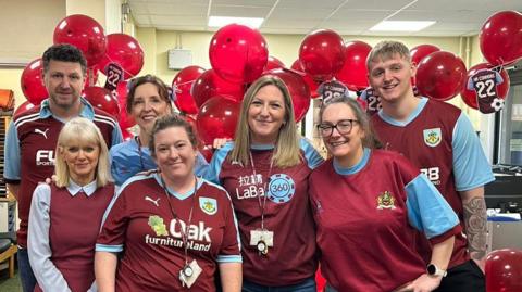 A mixed group of men and women wear a range of Burnley United Football Club's shirts - dark red body, light blue sleeves - or red and blue clothing. They stand in front of a group of red balloons, and bunting of small Burnley United football shirts.