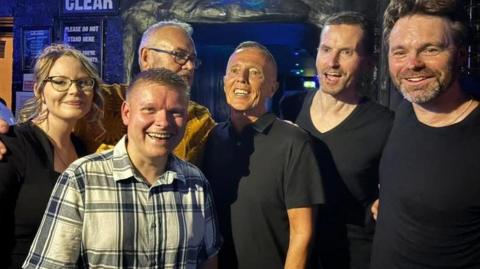 Photo of a band of five members and their idol, Curt Smith from Tears for Fears, standing with them in the middle. They're inside a bar and all look joyful.