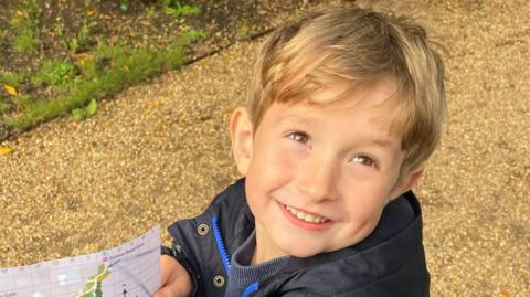 Benedict, a young boy with blond hair, on a path holding a map. He is wearing a black coat and is smiling to the camera.