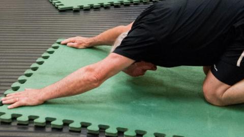 A man with grey hair is is stretching in "extended child pose" as it is called in yoga. He is on his knees, leaning forward with his arms outstretched and his forehead is nearly touching the green gym mat.