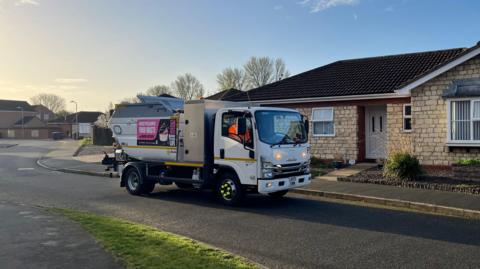 A small waste collection lorry with its lights on drives down a residential street at dawn.