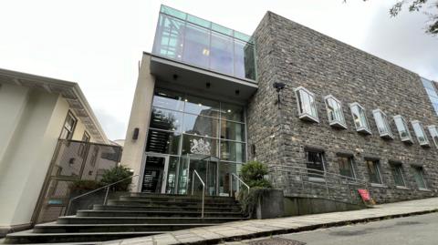 Guernsey’s Royal Court building with modern glass balconies and traditional stone facade rises beside a sloping street, featuring steps, railings, tall windows, nearby trees, and bright daylight urban surroundings quiet.