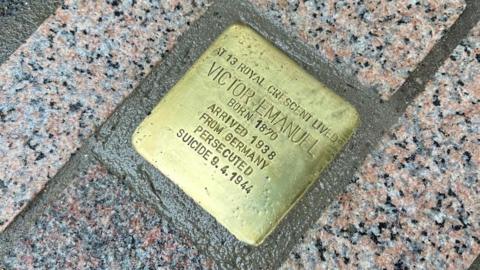 A picture of a brass plaque surrounded by stone on the floor. It has writing on it which states the name Victor Emanuel. 