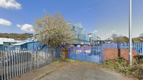 An industrial area. From a public footpath, you can see a warehouse building which houses the ironworks factory. The metal gates onto the ground are painted blue.