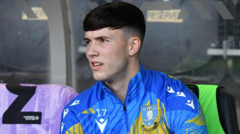 Charlie McNeill sits in the dugout during a game for Sheffield Wednesday. He is wearing a light blue and yellow club tracksuit top.