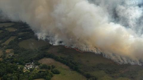 An aerial view of the fire shows an expanse of green fields with a line of fire and huge plumes of white smoke wafting up into the sky. In the left corner of the image, there is a plot of land which has some farm buildings on it, surrounded by green hedging.