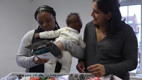 Rosabel is holding her baby over one shoulder whilst looking at the contents of the baby box. She is standing next to Roma Mascarenhas
who is the Family Hub Connector 
