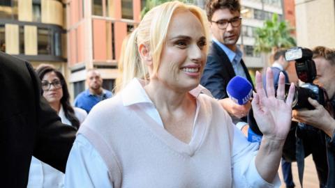 A woman waving at the camera outside court with blonde hair, wearing a white shirt and beige vest 