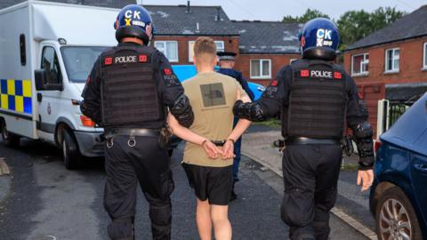 Two police officers in helmets and bullet-proof vests hold a man who is handcuffed around each arm and lead him towards a police van parked on the street. 