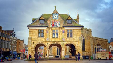 A historic-looking beige building. The downstairs is open with an enclosed upstairs. Several people are walking nearby and there is a row of shops to the left. A church is behind it and to the right.